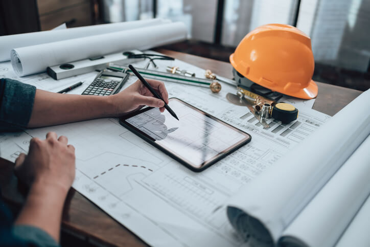 A man using the best estimating software for small contractors on an iPad on their desk, with construction blueprints and a hardhat also on the table.